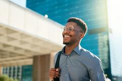 Homem jovem e sorridente andando ao ar livre em uma área urbana, com camisa social e mochila nas costas, representando confiança e entusiasmo no início da jornada profissional.