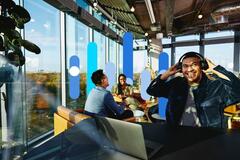 Colaboradores de diferentes origens interagindo em um escritório moderno com elementos gráficos em azul. Um profissional sorri com fones de ouvido enquanto outros conversam ao fundo. A imagem representa o impacto positivo da consultoria de diversidade e inclusão na construção de culturas corporativas mais representativas e colaborativas.