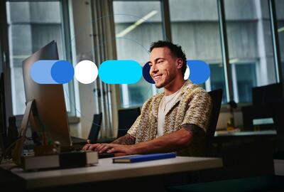 Homem vestido casualmente e utilizando computador em escritório, sorridente ao preencher habilidades profissionais no currículo.