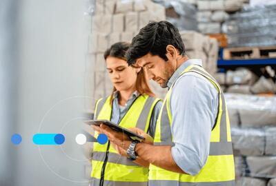 Homem e mulher em armazém de fábrica, vestindo roupas de segurança e pesquisando como e onde contratar profissional de logística.