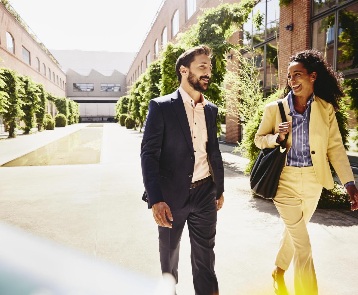 Man and woman talking and having a laugh while walking outside between office buildings.