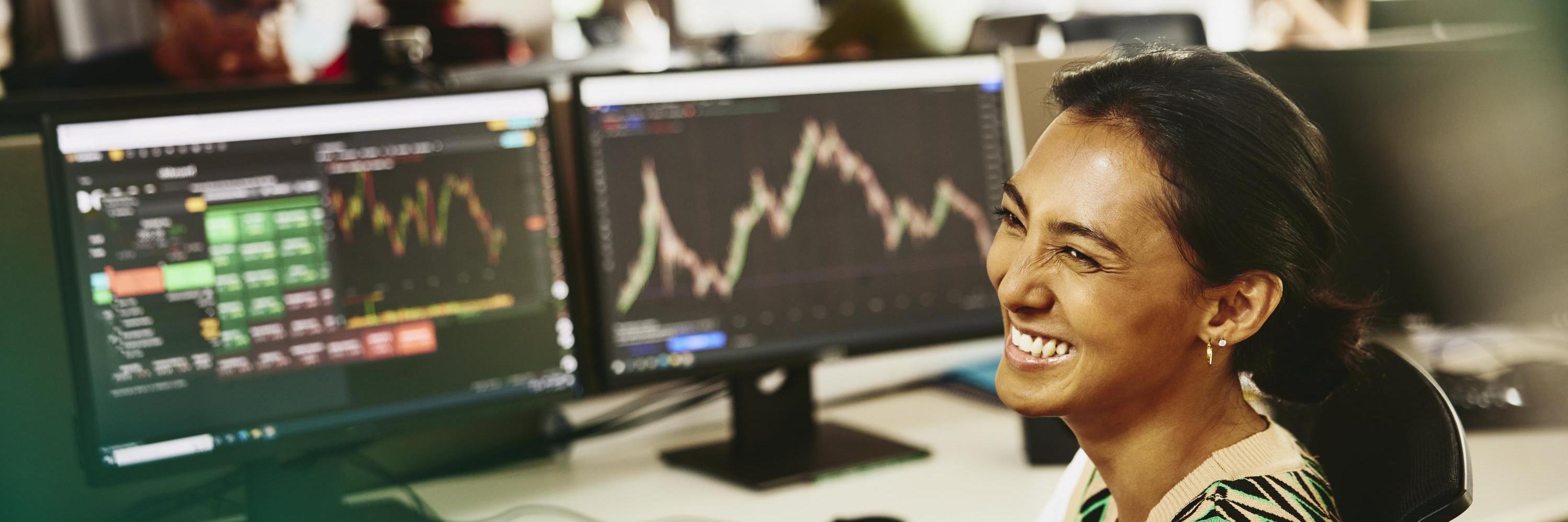 Woman smiling while sitting behind her desk, screens displaying financial information.
