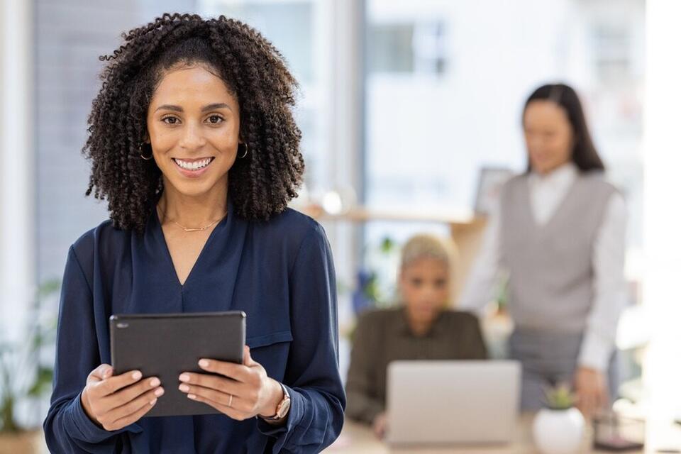 Mulher sorridente segurando um tablet em um ambiente de escritório, com colegas de trabalho ao fundo, ambiente corporativo moderno